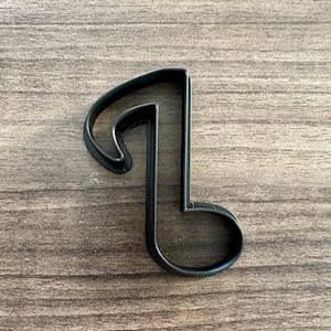 May include: A black, single eighth note cookie cutter. The cutter is made of plastic and is set against a wooden background. The musical note is a common symbol used in music and baking.