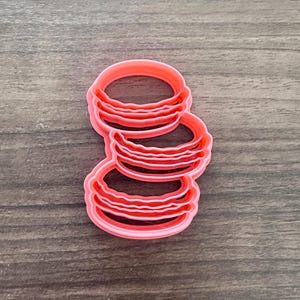 May include: A pink plastic cookie cutter in the shape of three stacked macarons. The cutter has a detailed design with ridged edges, perfect for creating decorative cookies. The cutter is on a wooden surface.