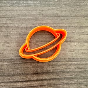 May include: An orange plastic cookie cutter in the shape of a planet with a ring around it. The cutter is made of a single piece of plastic and is designed to cut out the shape from cookie dough or other similar materials. The cutter is on a wooden surface.