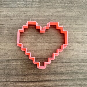 May include: A pink, pixelated heart-shaped cookie cutter. The cutter is made of plastic and has a geometric design, perfect for creating unique cookies. The cutter is displayed on a wooden surface.