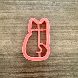 May include: A pink plastic cat-shaped cookie cutter. The cutter is a single piece with a simple outline of a sitting cat, including the head, body, and tail. The cutter is on a wooden surface.