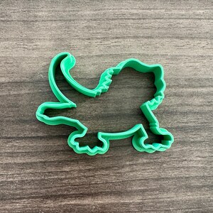 May include: A bright green plastic cookie cutter in the shape of a dinosaur. The cutter has a detailed outline, including the dinosaur's head, tail, and legs. The cutter is designed for making cookies or other treats.