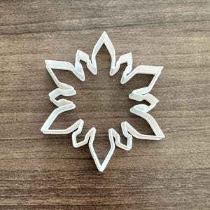 May include: A white plastic snowflake cookie cutter. The cutter is in the shape of a six-pointed star with intricate details. The cutter is on a brown wooden surface.