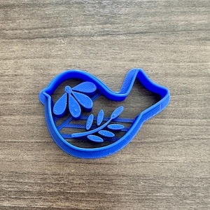May include: A blue plastic cookie cutter in the shape of a dove, with a floral design inside. The dove is facing left, with a flower and leaves inside its body. The cutter is made of a solid blue plastic.