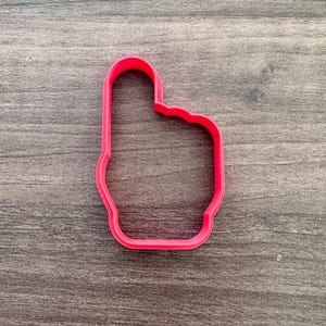 May include: A bright pink cookie cutter in the shape of a hand with the index finger pointing up. The cutter is made of plastic and sits on a wooden surface.