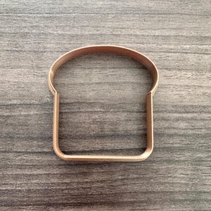 May include: A copper-colored metal cookie cutter in the shape of a slice of bread. The cutter has a textured edge and sits on a gray wooden surface. Ideal for making toast-shaped cookies or biscuits.