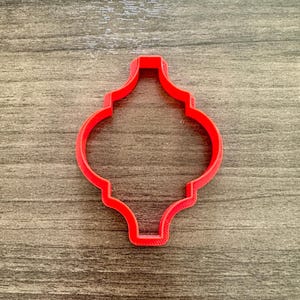 May include: A red plastic cookie cutter in a quatrefoil shape, ideal for baking. The cutter is made of a solid, bright red plastic and sits on a wooden surface.