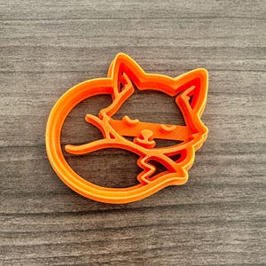 May include: Orange plastic cookie cutter in the shape of a curled-up fox. The cutter has a detailed design, showing the fox's head, body, and tail. The cutter is on a gray wooden surface.