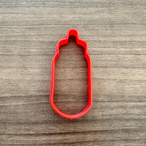 May include: A red plastic cookie cutter in the shape of a baby bottle. The cutter is a solid outline, with a rounded base and a nipple-shaped top. The cutter is on a wooden surface.