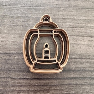 May include: A light brown lantern-shaped cookie cutter with a candle design inside. The cutter has a rounded top and bottom with a handle-like feature at the top. The design is a single piece of plastic.