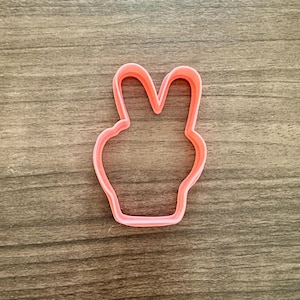 May include: A pink plastic cookie cutter in the shape of a peace sign, with the fingers raised. The cutter is set against a wooden background.