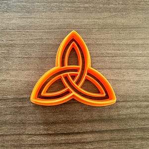 May include: An orange and black Celtic knot cookie cutter. The triquetra design is a symbol of interconnectedness. The cookie cutter is made of plastic and is suitable for making cookies or other baked goods. The cutter is approximately 3 inches wide.