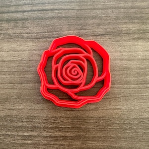 May include: A red plastic rose-shaped cookie cutter. The cutter is a single piece with a detailed outline of a rose, including petals and a central spiral. The cutter is designed for baking and crafting.