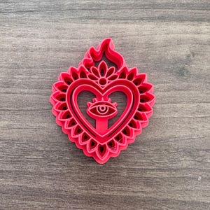 May include: A bright pink cookie cutter in the shape of a heart with an eye in the center. The heart is surrounded by a floral design and topped with a flame. The cutter is made of plastic and is on a wooden surface.