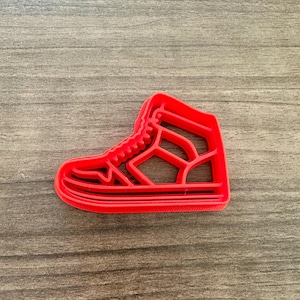 May include: A bright red cookie cutter in the shape of a high-top sneaker. The cutter has a detailed design, including the laces, sole, and side panels. The cutter is made of plastic and is designed for baking.