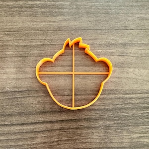 May include: An orange plastic cookie cutter in the shape of a lion's head. The cutter has a simple design with a cross-shaped interior support structure. The cutter is on a wooden surface.