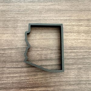 May include: A dark gray cookie cutter in the shape of the state of Arizona. The cutter is made of a solid material and has a rectangular shape with the state's outline cut out. The cutter is placed on a wooden surface.