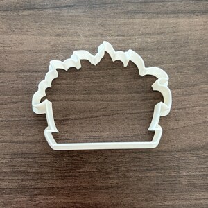 May include: A white plastic cookie cutter in the shape of a cupcake. The cutter has a scalloped top edge and a rectangular base. The cupcake design is a common theme for baking and crafting.