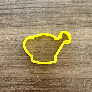 May include: A yellow plastic cookie cutter in the shape of a watering can. The cutter is a solid outline, with a handle and spout. The cutter is on a brown wooden surface.