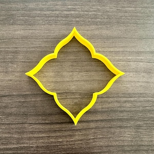 May include: A yellow, four-lobed cookie cutter with a decorative, symmetrical design. The cutter is made of plastic and is set against a wooden background.