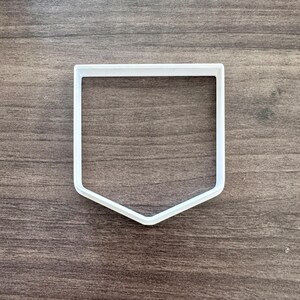 May include: A white plastic cookie cutter in the shape of a pocket. The cutter has a rectangular top with a rounded bottom. The cutter is on a brown wooden surface.