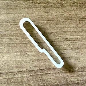 May include: A white, hollow, plastic-like outline shaped like a simple kitchen knife. The object features a long, narrow handle section that transitions into a wider blade area, forming a continuous, thin, rounded frame with an open center.