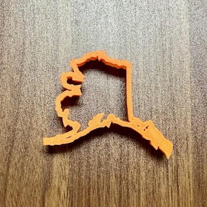 May include: An orange plastic cookie cutter shaped like the state of Alaska.