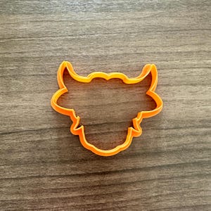 May include: An orange plastic cookie cutter in the shape of a bull's head. The cutter has a detailed outline of the bull's horns, ears, and face. The cutter is placed on a wooden surface.