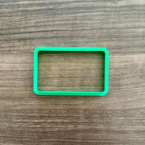 May include: A bright green rectangular cookie cutter made of plastic. The cutter has rounded corners and is set against a wooden surface. The cookie cutter is a simple shape, perfect for making cookies or other treats.