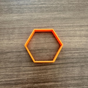 May include: An orange, hexagon-shaped cookie cutter. The cutter is made of plastic and sits on a wooden surface. The cutter is a bright orange color and has six sides.