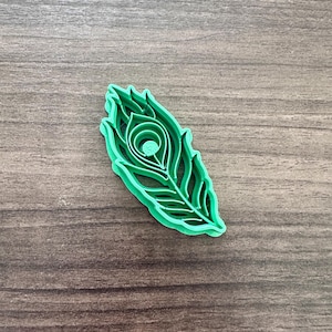 May include: A bright green peacock feather-shaped cookie cutter. The cutter is made of plastic and features a detailed design of a peacock feather, including the eye and the feathery edges. The cutter is on a wooden surface.