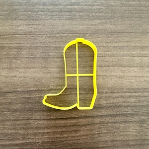 May include: A yellow plastic cookie cutter in the shape of a cowboy boot. The boot has a pointed toe, a tall shaft, and a small heel. The cutter is on a wooden surface.