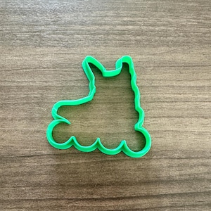 May include: A bright green llama-shaped cookie cutter. The cutter is made of plastic and has a simple outline of a llama with a curved base. The cutter is placed on a wooden surface.