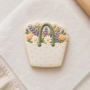 May include: A decorated sugar cookie shaped like a tote bag filled with flowers. The bag is off-white with gray polka dots and a green handle. The flowers include daisies, lavender, and tulips in shades of yellow, orange, and purple. The cookie is on white parchment paper.