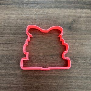 May include: A bright pink cookie cutter in the shape of a bow with a face. The cutter has a rectangular base and a scalloped edge on the sides. The bow is at the top of the cutter.