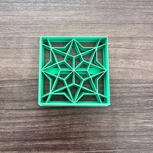 May include: A square, bright green cookie cutter with a geometric starburst design. The cutter features a series of interconnected star shapes within a square frame. The cutter is made of plastic and sits on a wooden surface.