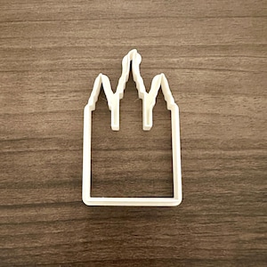 May include: A white plastic cookie cutter in the shape of a castle or building with a pointed roof and rectangular base. The cutter has a simple, clean design, perfect for baking themed treats.