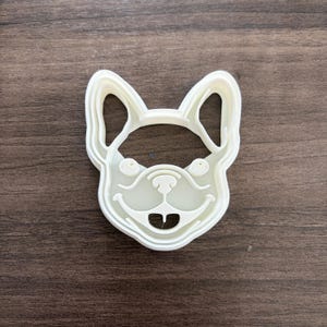 May include: A white plastic cookie cutter in the shape of a French Bulldog's face. The cutter features the dog's ears, eyes, nose, and mouth, with a smiling expression. The cutter is designed for making cookies or other treats.