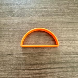 May include: An orange, semi-circular cookie cutter. The cutter is made of plastic and has a flat bottom with a curved top. The cutter is on a wooden surface.