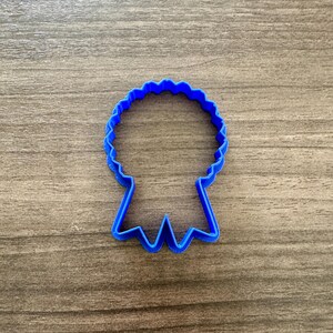 May include: A blue plastic cookie cutter in the shape of a ribbon. The cutter has a scalloped top and two ribbon tails. The cutter is on a wooden surface.