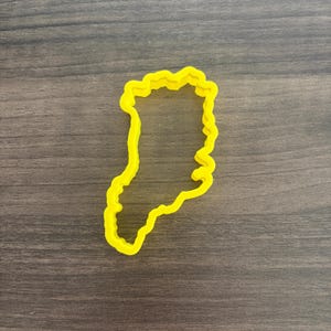 May include: A bright yellow cookie cutter in the shape of a geographical outline. The cutter has a wavy edge and is made of plastic. The cutter is on a dark wood grain surface.