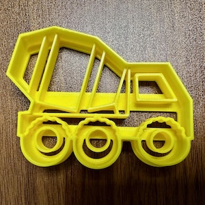 May include: A bright yellow cookie cutter in the shape of a dump truck. The cutter has a detailed design with a cab, bed, and three wheels. The cutter is made of plastic and is designed for baking.