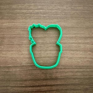 May include: A bright green cookie cutter in the shape of a potted plant. The cutter is made of plastic and has a simple, clean design. The cutter is placed on a wooden surface.