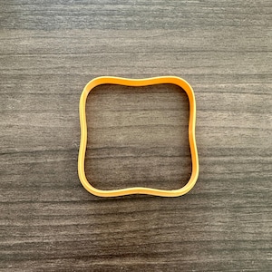 May include: An orange, square-shaped cookie cutter with slightly curved sides. The cutter is made of plastic and sits on a dark wood-grain surface. The cutter is designed for baking and crafting.