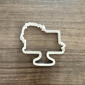 May include: A white plastic cookie cutter in the shape of a cake. The cutter has a rectangular top with a scalloped edge, supported by a pedestal base. The design is simple and clean, perfect for baking.