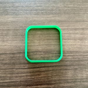 May include: A bright green, square-shaped cookie cutter with rounded corners. The cutter is made of plastic and sits on a wooden surface. The design is simple and clean, perfect for baking.
