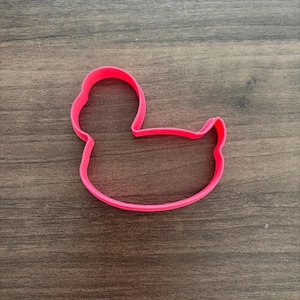 May include: A bright pink cookie cutter shaped like a duck. The outline features a rounded head, a curved neck, a smooth back, and a small tail section. The frame is constructed from a single continuous piece of molded plastic with a flat edge.