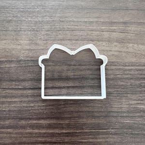 May include: A white plastic cookie cutter in the shape of a gift box with a bow on top. The cutter has a rectangular base and a curved top, perfect for creating festive treats. The cutter is shown on a wooden surface.