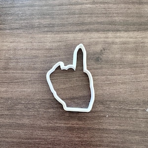 May include: White plastic cookie cutter in the shape of a hand with the middle finger extended. The cookie cutter is on a brown wooden surface.