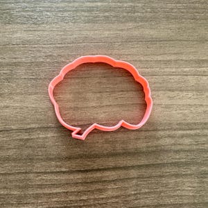 May include: A pink plastic cookie cutter in the shape of a thought bubble. The cutter is on a wooden surface. The cookie cutter is a solid color and has a smooth finish. The design is a simple outline.